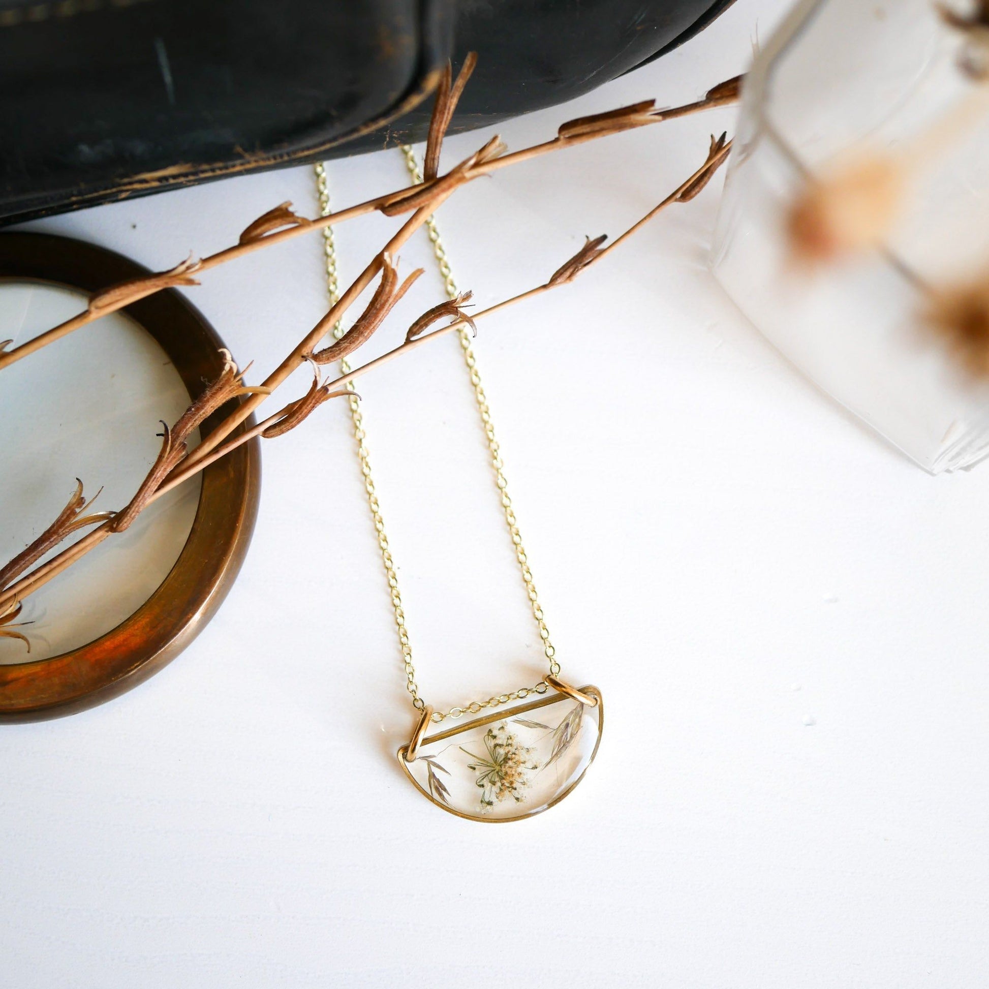 Flat lay of Meadow Luna Necklace with gold chain and botanical resin pendant styled with dried plants and vintage decor.