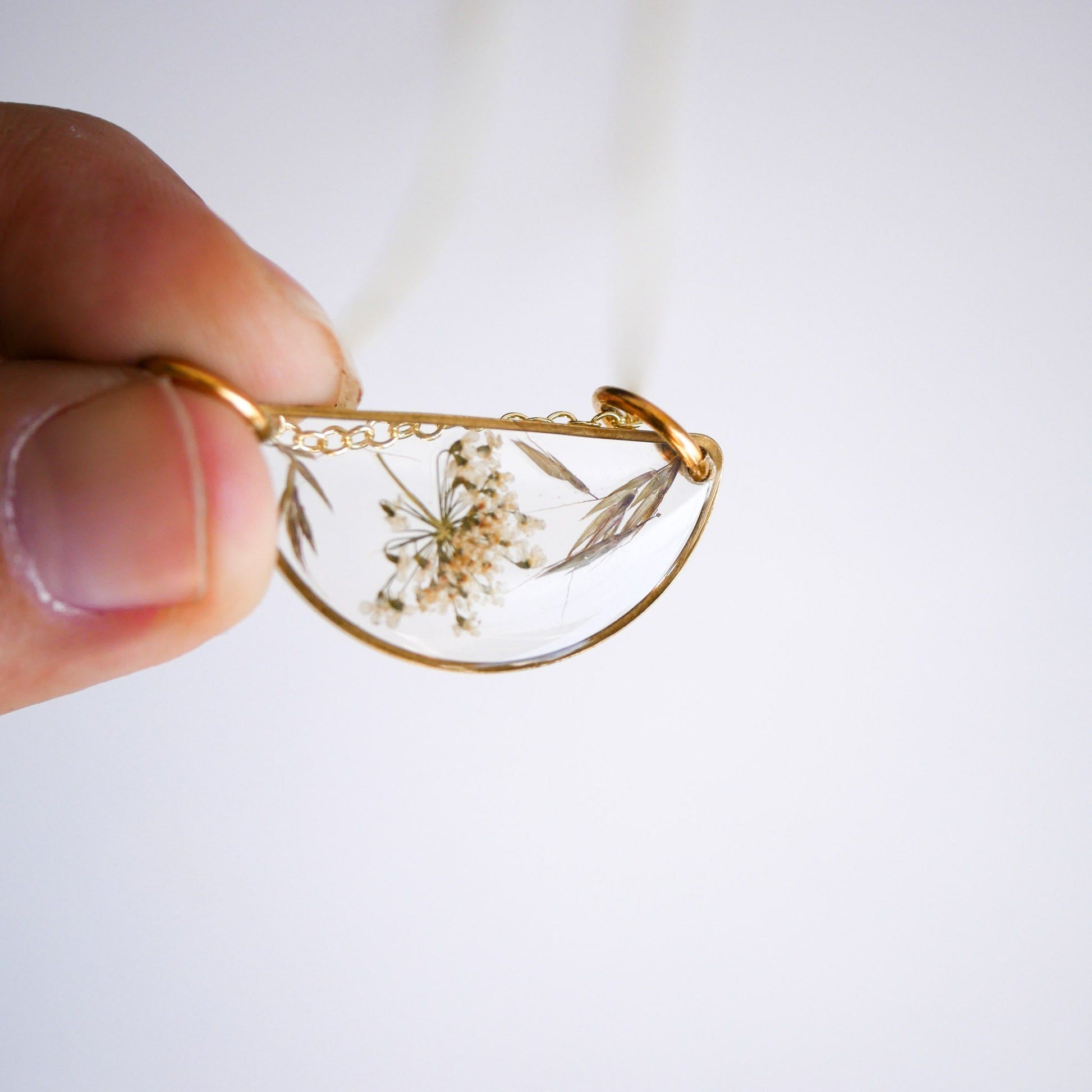 Hand holding the Meadow Luna Necklace showcasing its delicate botanical details in a transparent resin pendant with gold-plated edges.