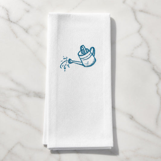 Uni-T white flour sack kitchen towel featuring a blue embroidered watering can design on a marble surface.