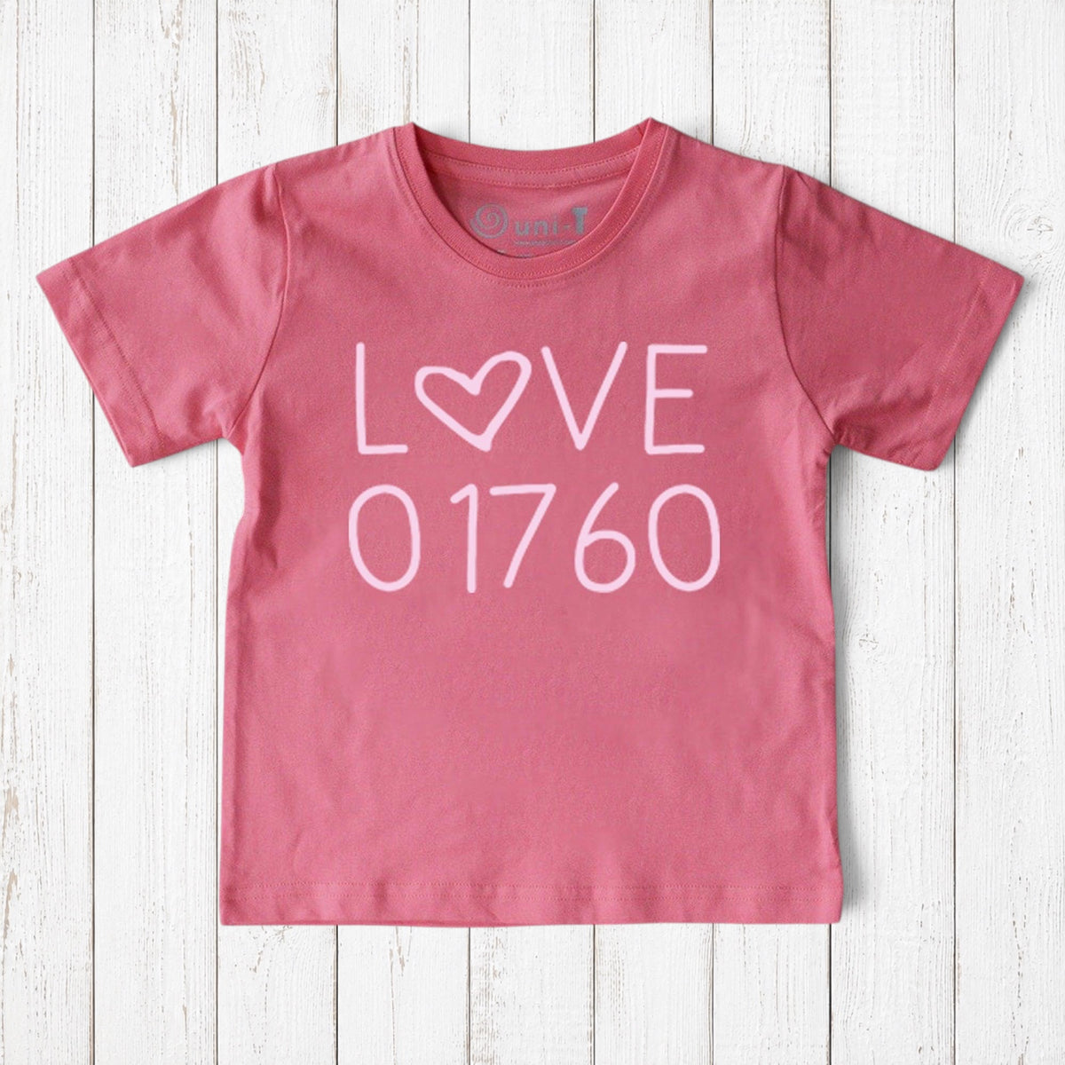 Uni-T kids pink organic cotton t-shirt featuring a white Love 01760 graphic print on a rustic wooden background.