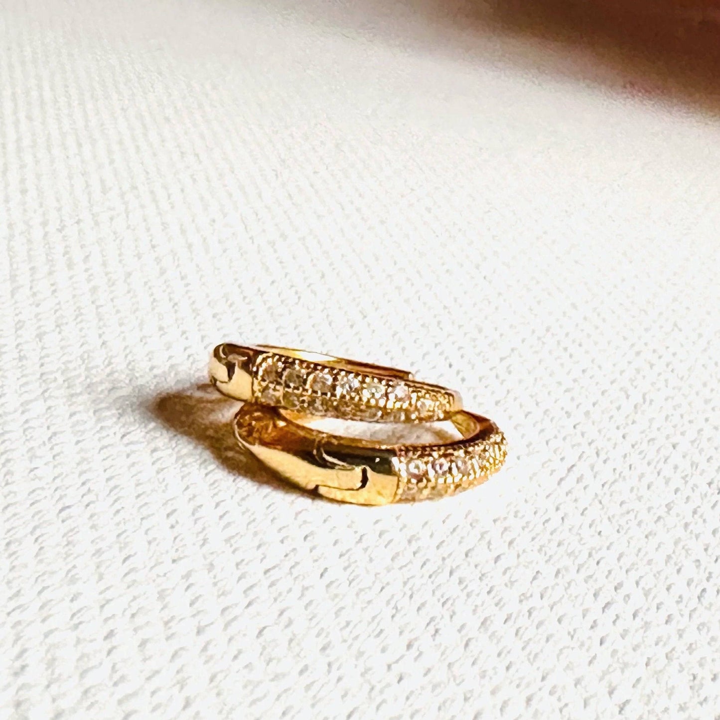 Gold huggie hoop earrings with sparkling pavé stones, resting on fabric. Secure hinged closures for comfort and elegance.