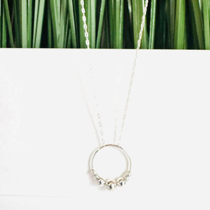 Silver Circle Necklace displayed against a green and white background. Features spinning sterling beads on a delicate 18” chain.