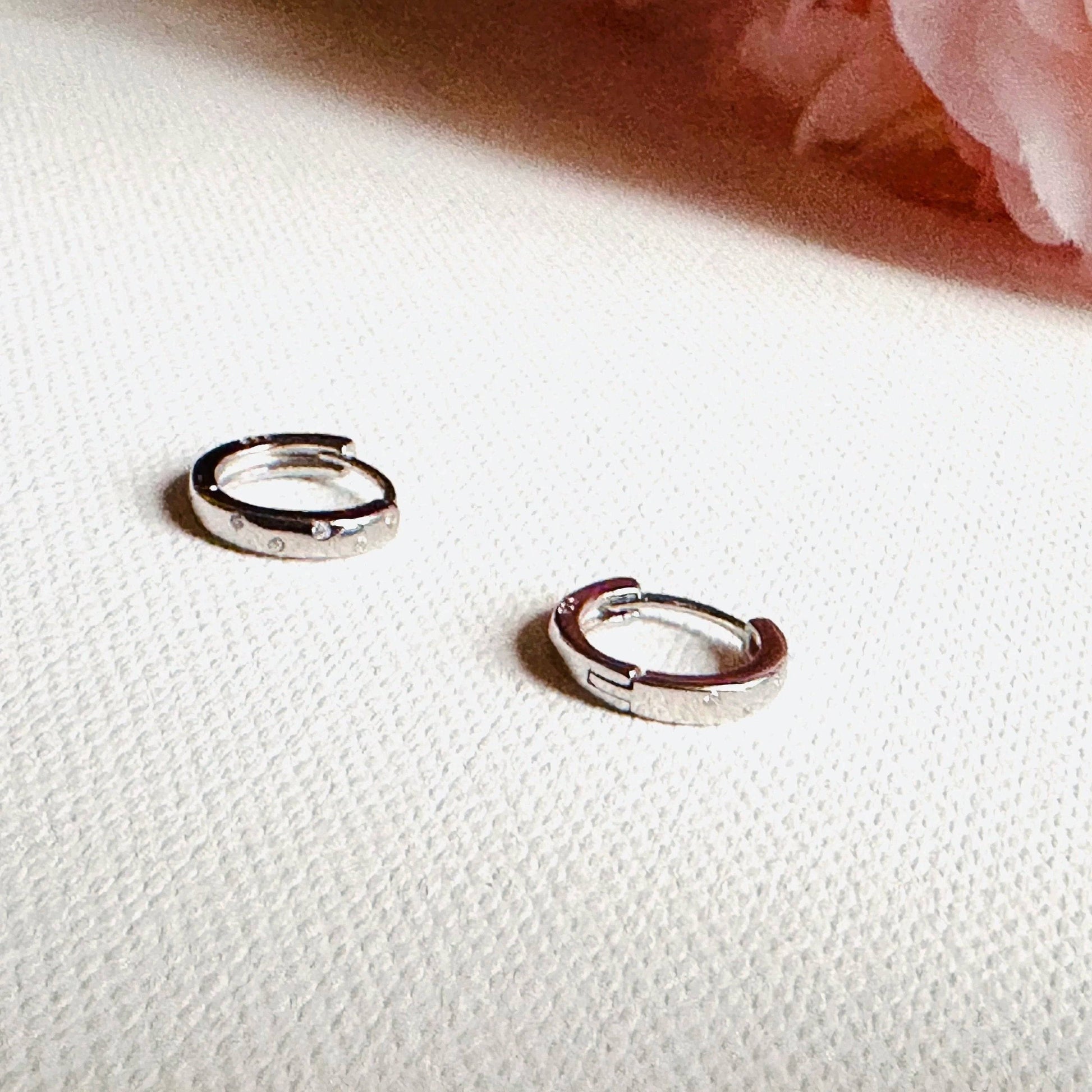 Pair of silver huggie hoop earrings with hinged closures on a soft textured surface. Timeless design for daily wear.