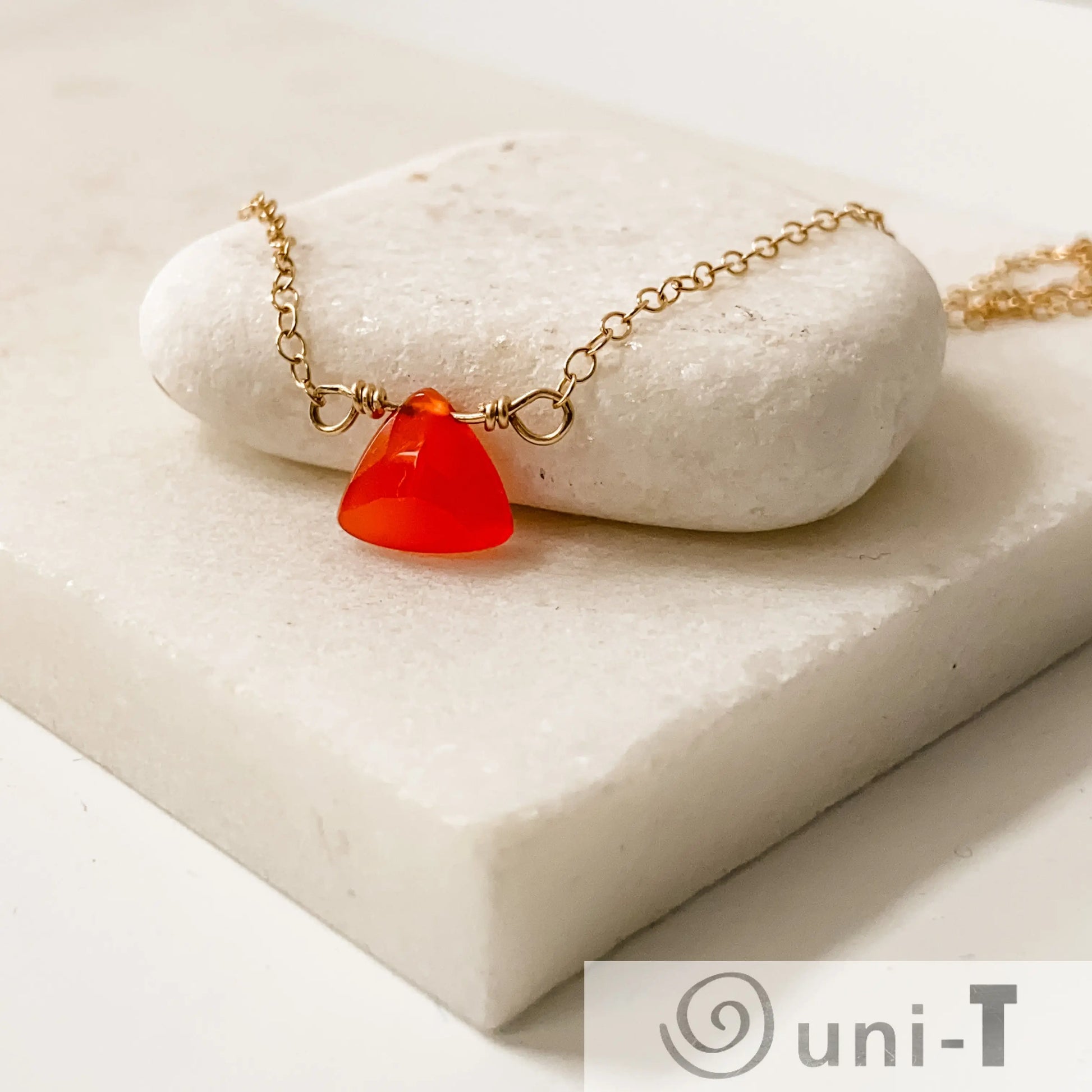 Carnelian Gemstone Necklace by Uni-T featuring a triangular red gemstone pendant on a gold-filled chain resting on white stone slab.