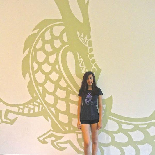 Model posing in front of a large dragon mural while wearing the 'Dare More' bamboo-organic cotton T-shirt. Eco-friendly mythical creature tee by Uni-T.
