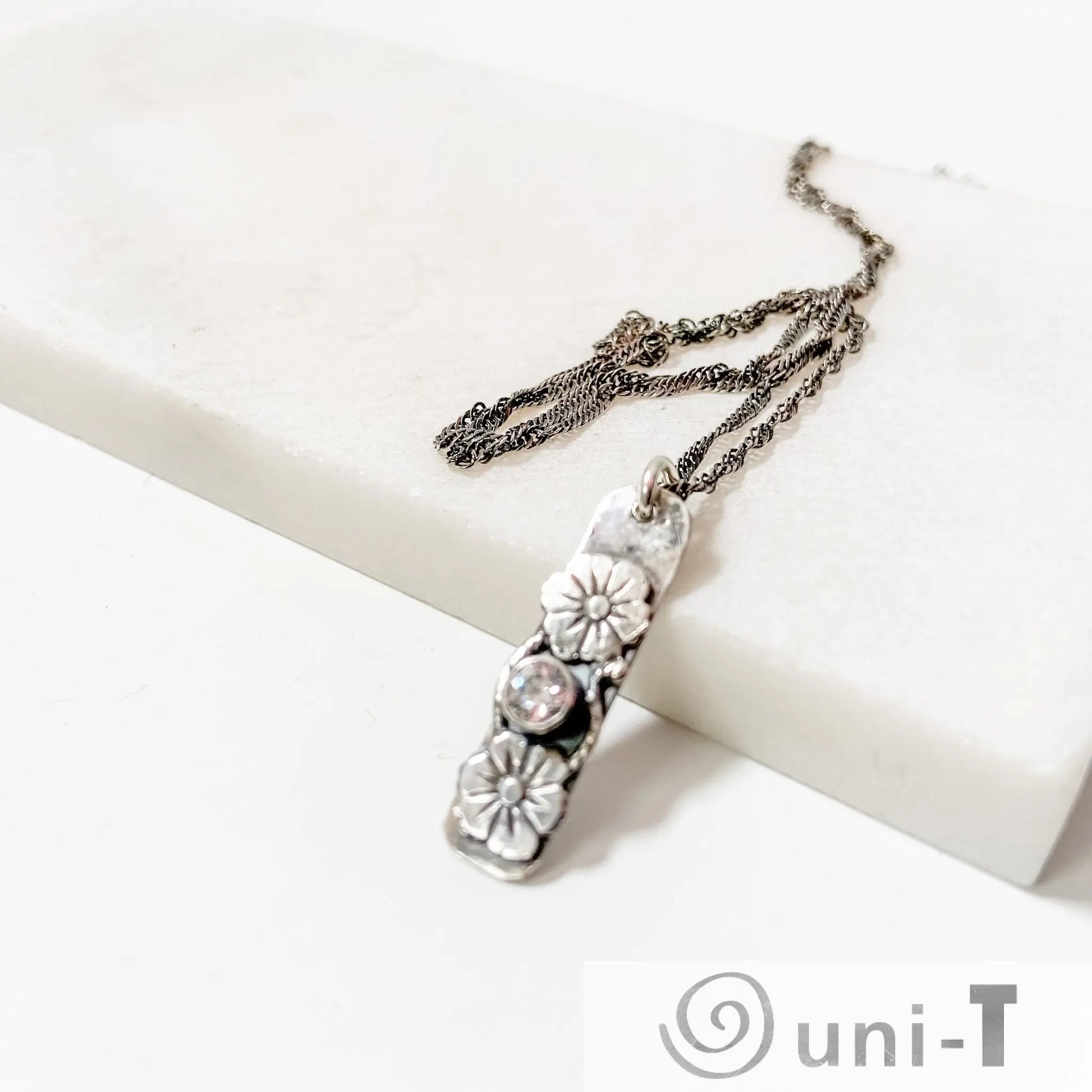 Flower Tag Necklace with cubic zirconia and floral silver tag pendant, displayed on a white marble surface with twisted rope chain.