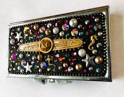 Round brass pill box with black velvet top, adorned with gold accents, pearls, rhinestones, and sea-themed charms for jewelry or makeup storage.