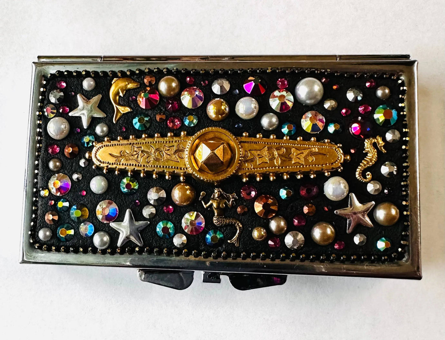 Handcrafted round brass pill box featuring a mosaic of colorful rhinestones, pearls, and metallic sea motifs on a black background.