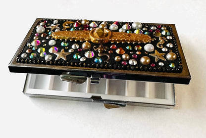 Open round brass pill box with six compartments, decorated with multicolor gems and gold embellishments, ideal for storing pills, jewelry, or makeup.