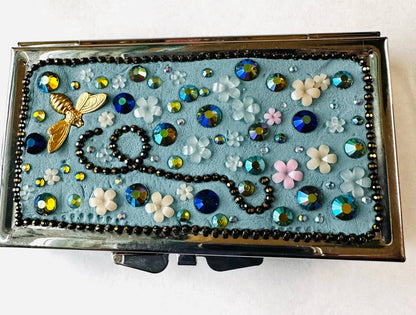 Blue-themed round brass pill box with floral resin decorations, rhinestones, and a gold bee charm, perfect for makeup or jewelry storage.