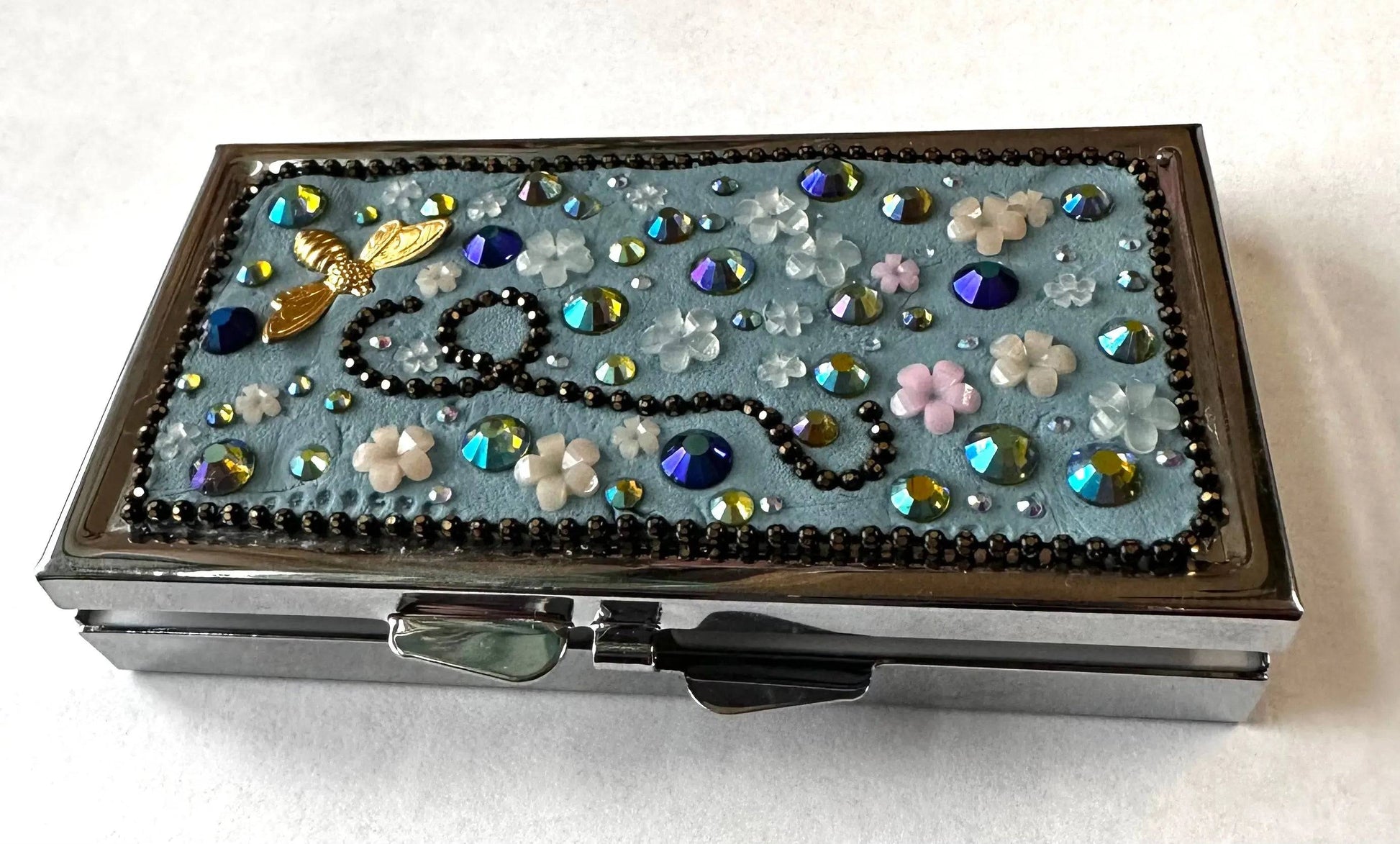 Compact round brass pill box with a blue floral mosaic top, featuring rhinestones and a bee charm, ideal for gifting or personal use.