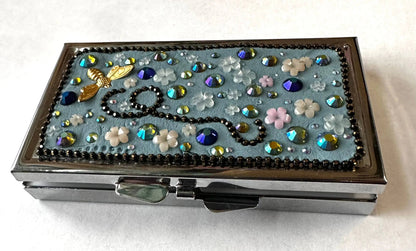 Compact round brass pill box with a blue floral mosaic top, featuring rhinestones and a bee charm, ideal for gifting or personal use.