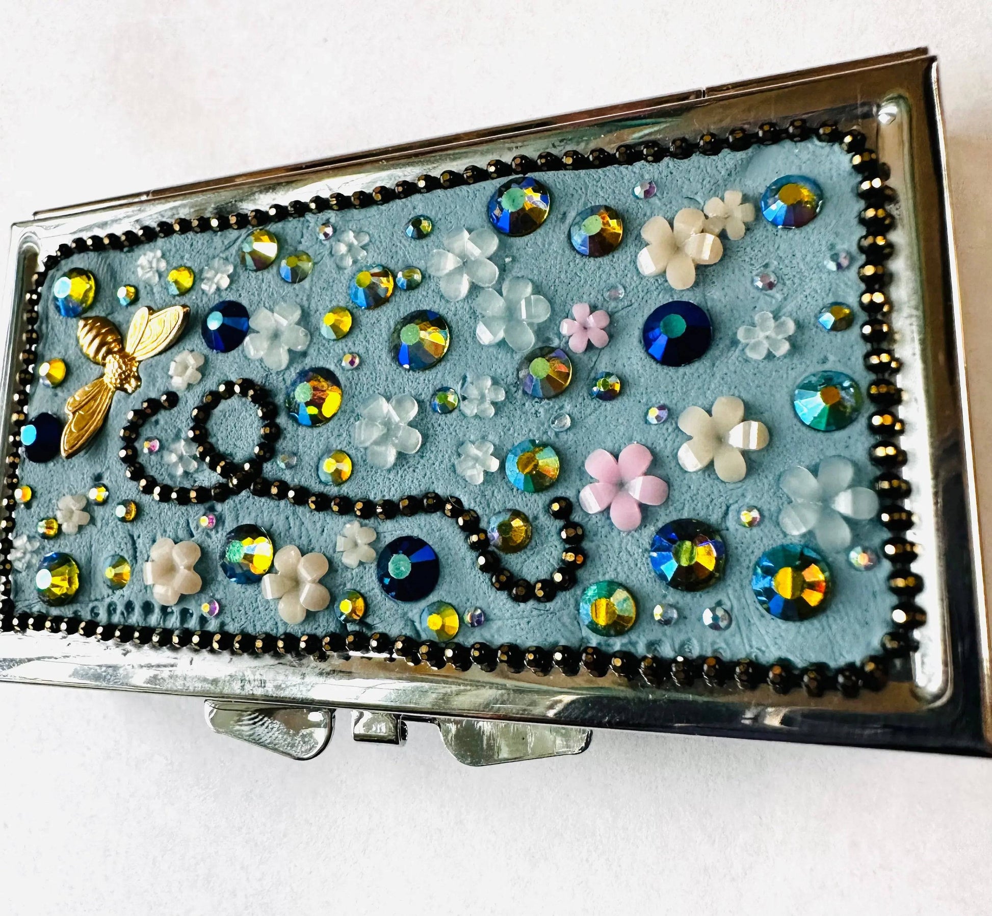 Decorative round brass pill box with blue background, floral accents, and sparkling rhinestones, designed for storing pills, jewelry, or makeup.