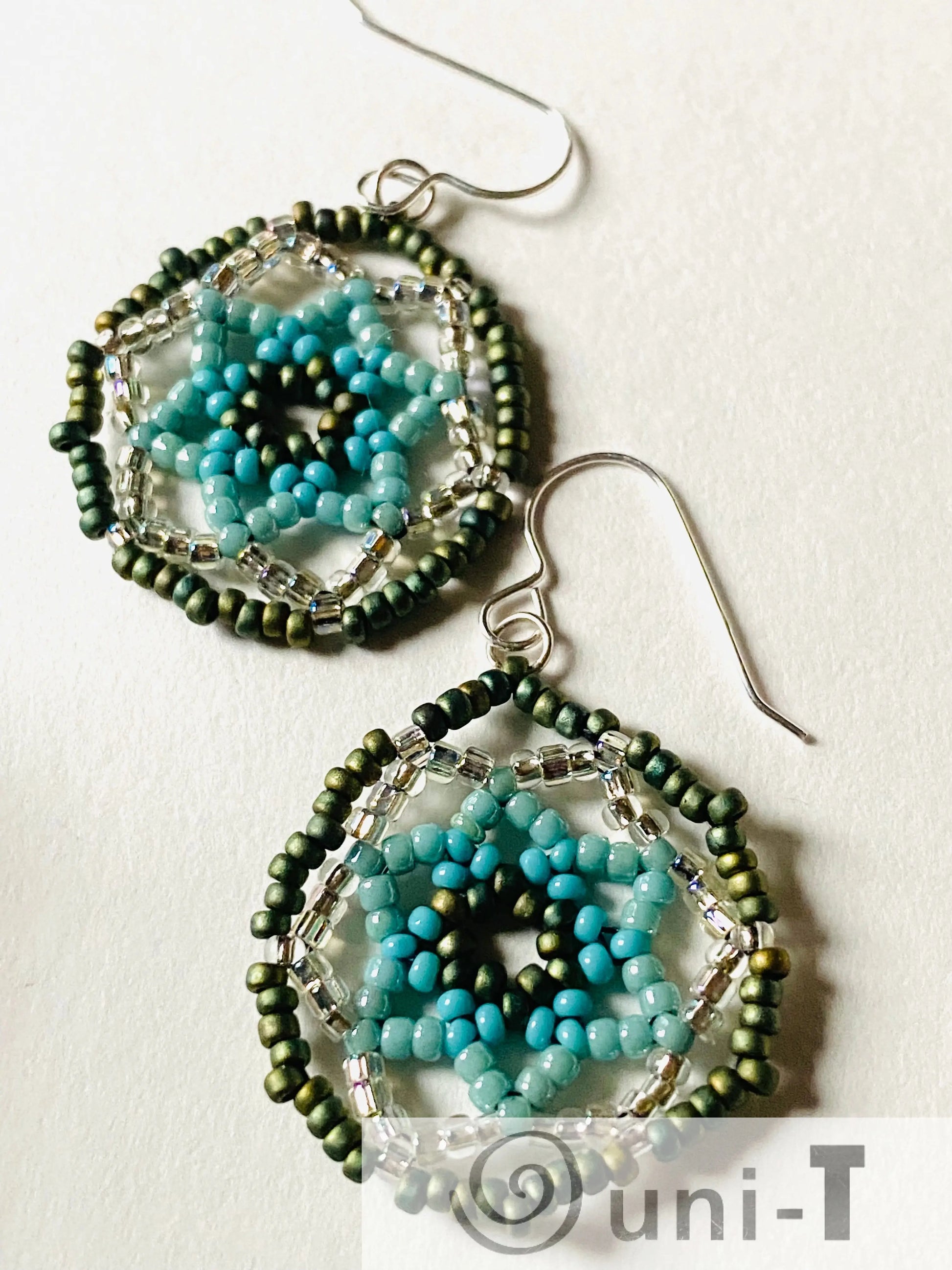Close-up of seafoam green medallion earrings with detailed blue and green beaded flower design and silver ear wires, highlighting craftsmanship.