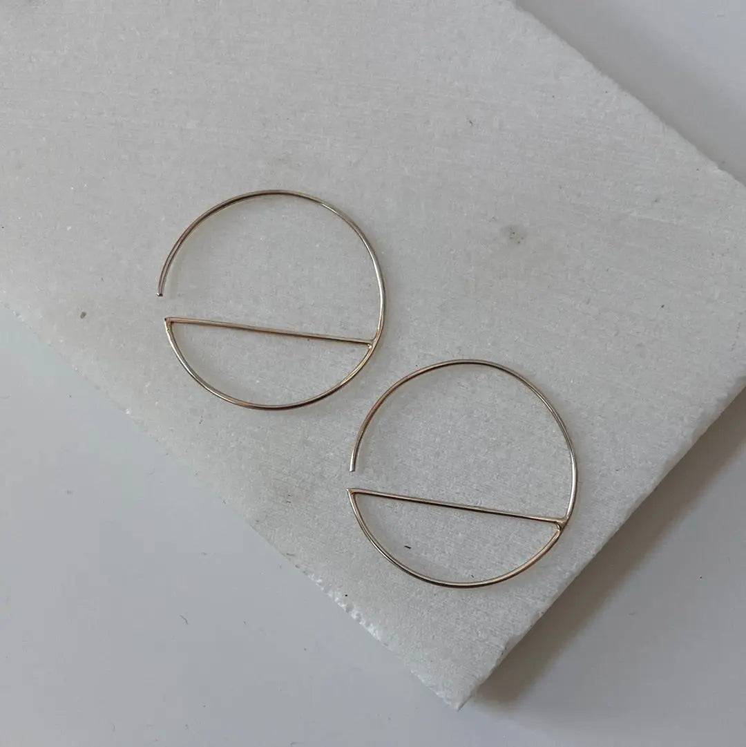 Flat lay of modern silver hoops with half-circle bar design. Sterling silver earrings with 1” diameter and clean, elegant lines.