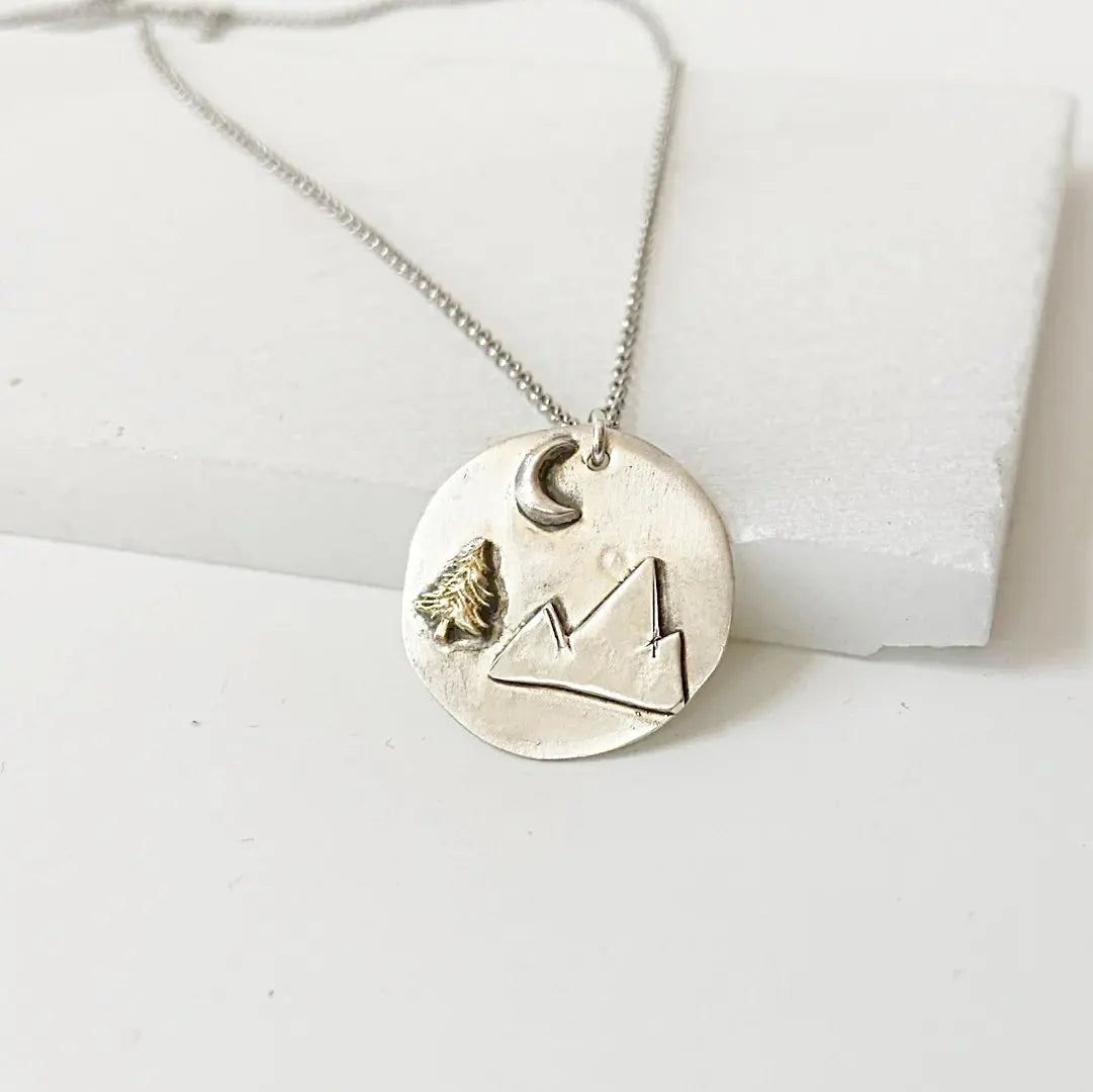 Mountain Medallion Necklace with raised mountain design, brass tree accent, and crescent moon on a silver-toned disc pendant and antiqued chain.