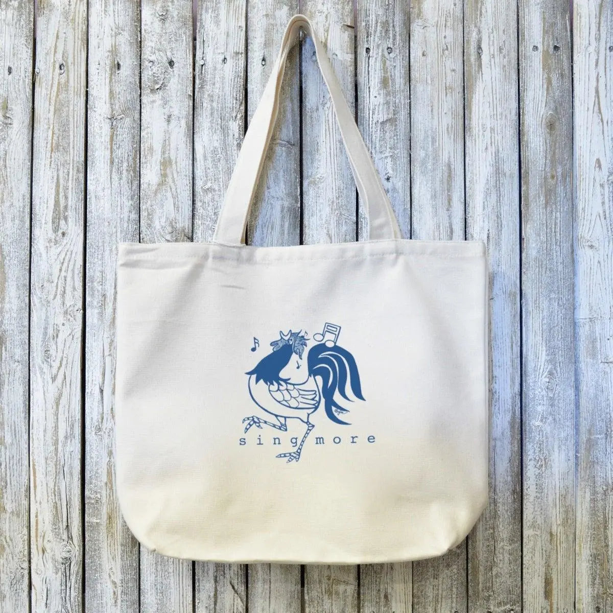 Organic canvas tote bag for singers featuring a blue hand-drawn rooster with musical notes and 'sing more' text. Eco-friendly and artist-designed.