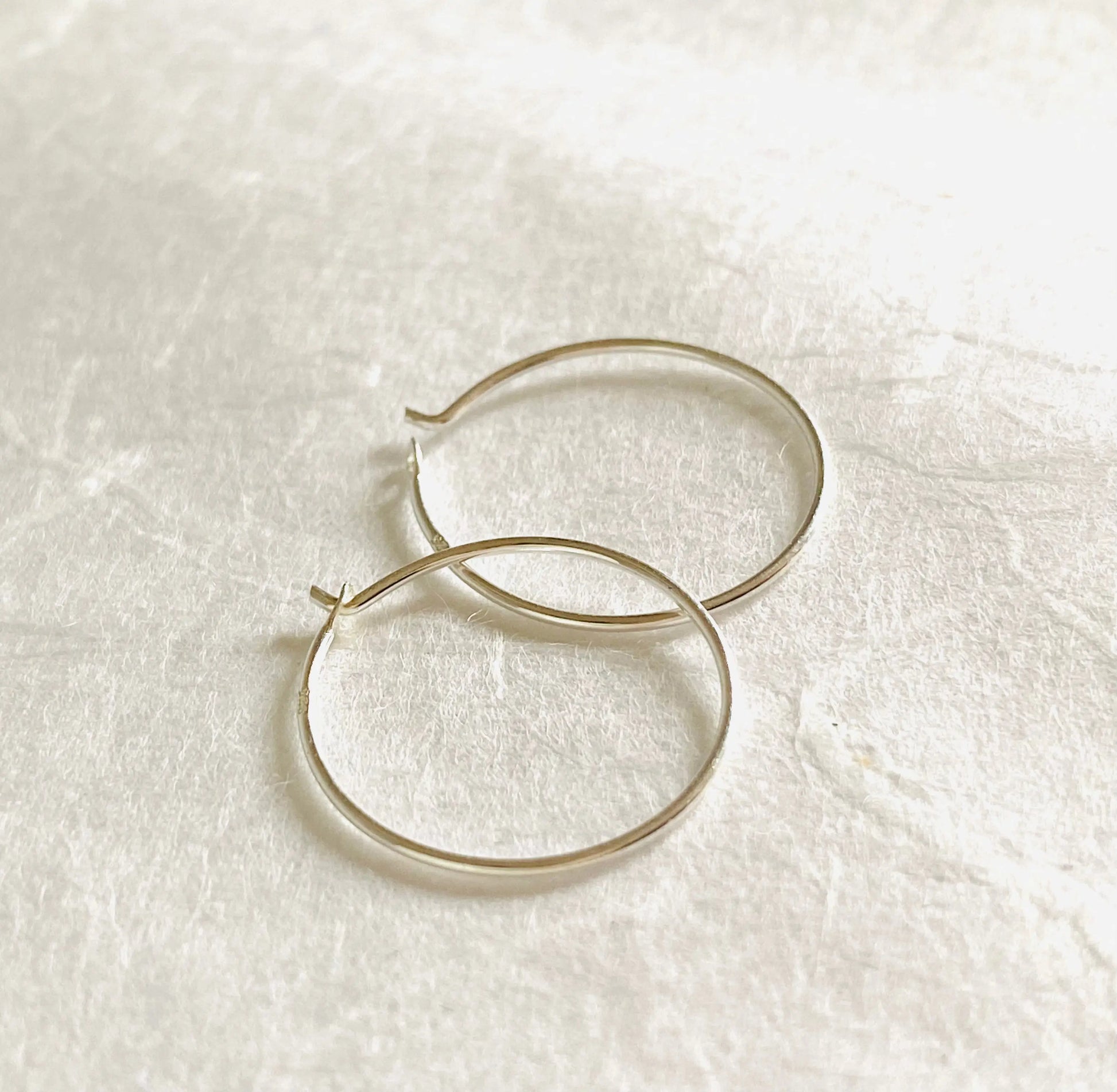 Minimalist 3/4” sterling silver hoop earrings on textured white surface. Elegant and lightweight hoops crafted from 22 gauge silver wire.