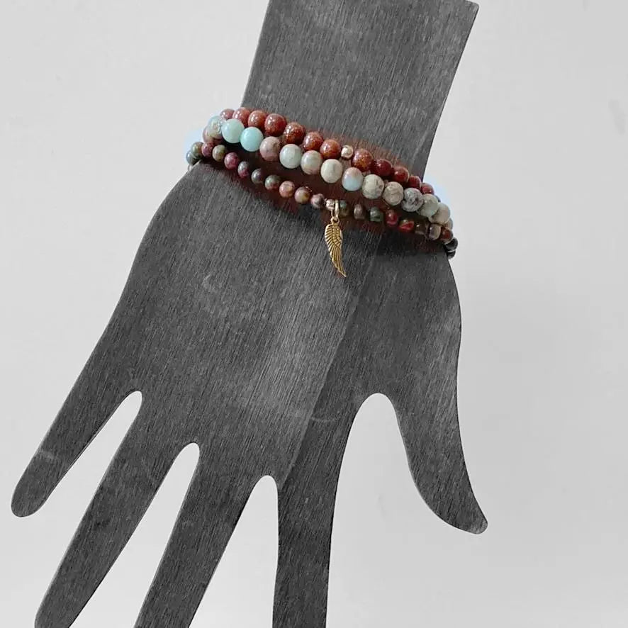 Triple Bracelet Stack worn on display hand, showcasing Ocean Jasper, Goldstone, and forest jasper beads with a brass wing charm accent.