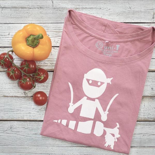 Folded pink 'Veggie Ninja' vegan T-shirt for women with white ninja and carrot graphic, made from bamboo and organic cotton. Displayed with fresh vegetables.